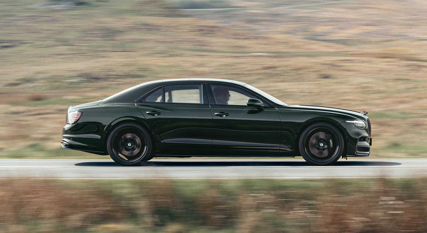 A sleek black sedan drives swiftly on a rural road, with blurred fields in the background, conveying a sense of speed and elegance.